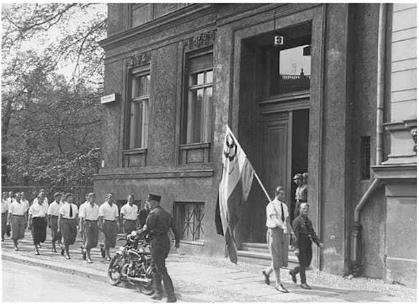 A black and white photograph of a student Nazi parade marching in front of the Institute for Sexual Sciences. Original caption: Students organized by the Nazi Party parade in front of the building of the Institute for Sexual Research in Berlin prior to pillaging it on May 6, 1933. United States Holocaust Memorial Museum