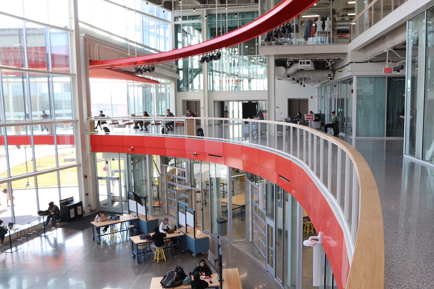 Maker spaces surround a brightly daylighted curving atrium. The glass walls of a theater can be seen beyond.