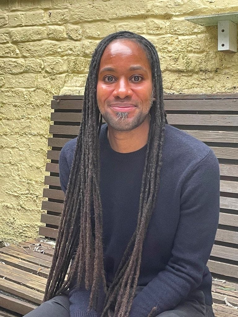 Jason Arday, a Black man with dreadlocks and a light beard, smiles while sitting on a wooden bench against a yellow brick wall. Jason Arday, a Black man with dreadlocks and a light beard, smiles while sitting on a wooden bench against a yellow brick wall.