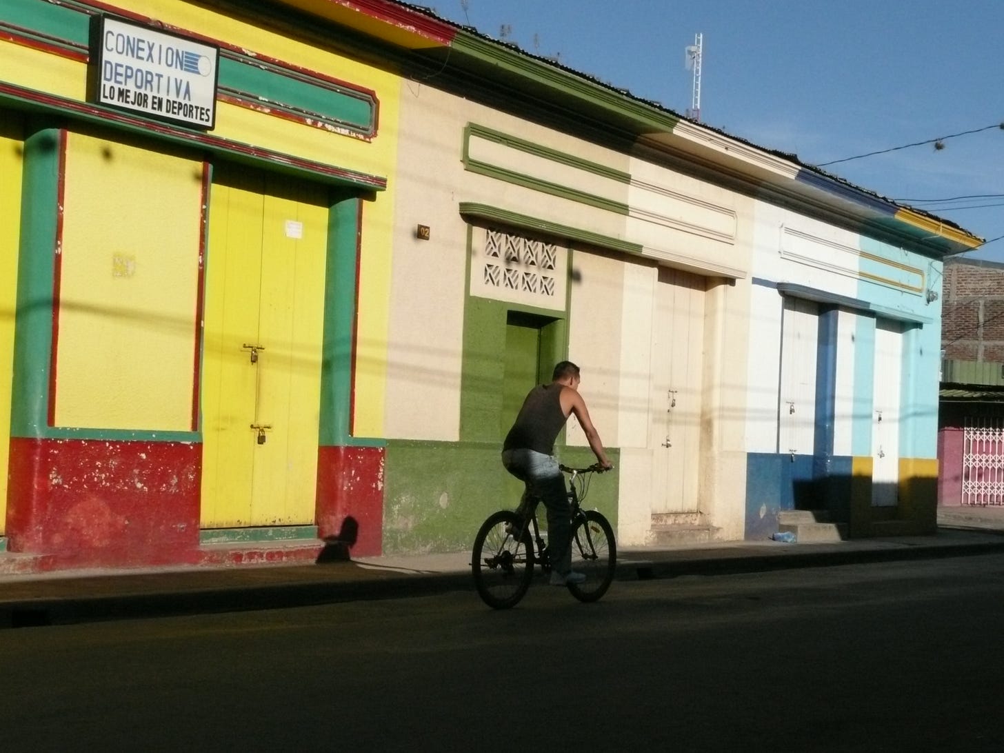 An image of a quiet street with a cyclist going past - he is wearing a black top and is positioned in the middle of the frame, surrounded by three different coloured one-storey buildings. The buildings doors are all closer=d. The sky is blue in the top corner of the picture. 