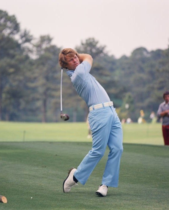 This may contain: a man swinging a golf club on top of a lush green field with trees in the background This may contain: a man swinging a golf club on top of a lush green field with trees in the background