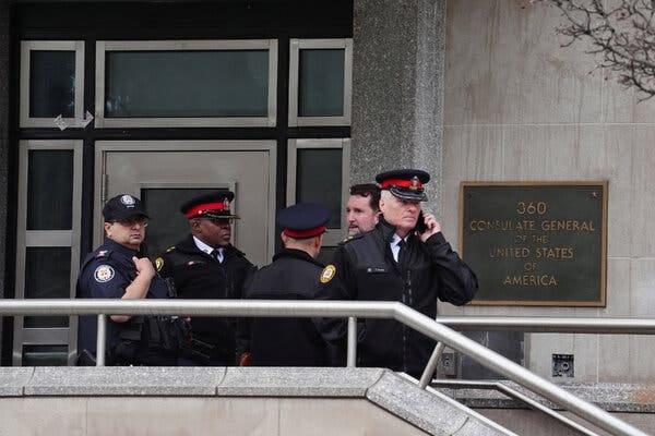 Several people in black uniforms with hats stand in front of a building with a sign that reads, “Consulate General of the United States of America.” One person is speaking on a phone. Several people in black uniforms with hats stand in front of a building with a sign that reads, “Consulate General of the United States of America.” One person is speaking on a phone.