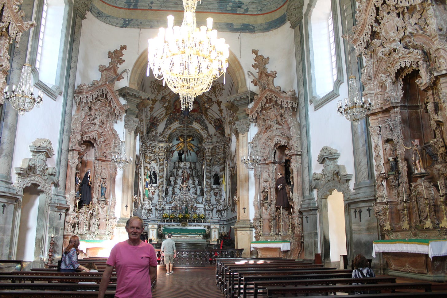 Foto do meu pai dentro de uma igreja bastante ornamentada de Minas. Ele veste camiseta rosa e sorri para a foto