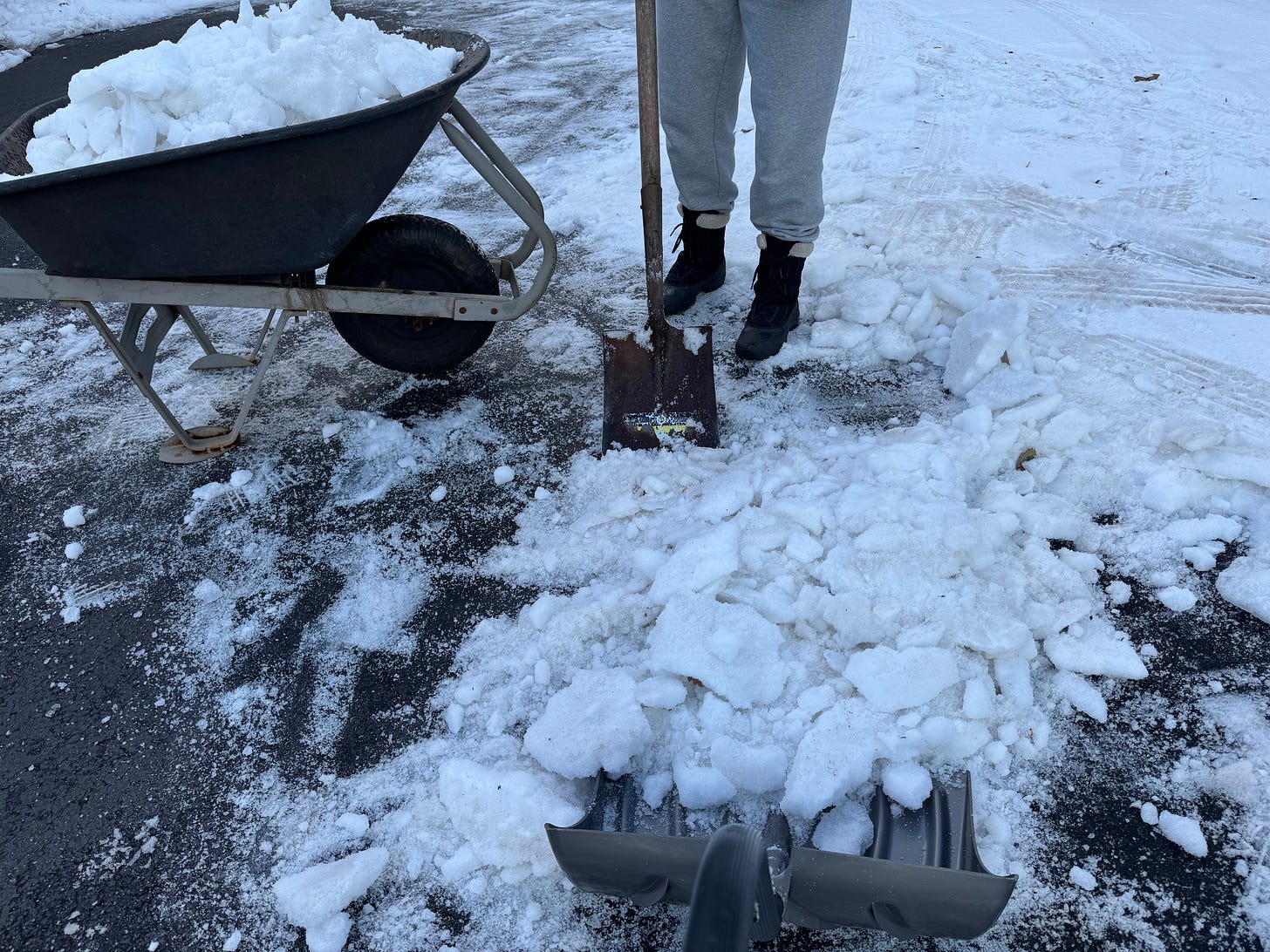 Two-person team removing a thick blanket of snow-ice from the driveway. One raises and breaks it with a metal garden shovel and the other loads it into a wheelbarrow with a large snow shovel. No faces shown.