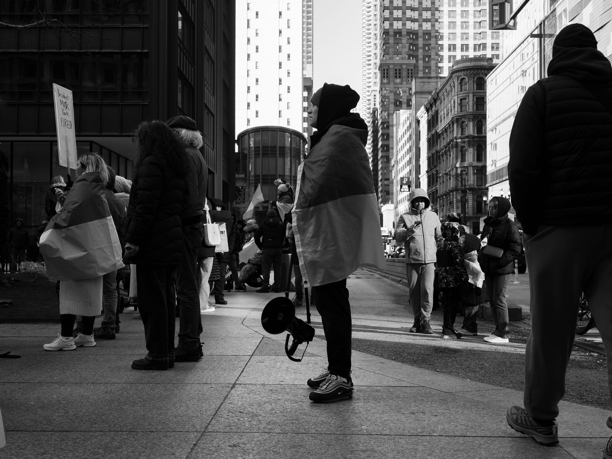 A black and white photograph captures a protest scene in an urban setting. A person draped in a Ukrainian flag stands in the foreground, holding a megaphone at their side. Other demonstrators, also wrapped in flags, engage in conversation or hold signs. The background features towering buildings and city streets, emphasizing the scale of the demonstration. The image conveys a moment of reflection and determination. A black and white photograph captures a protest scene in an urban setting. A person draped in a Ukrainian flag stands in the foreground, holding a megaphone at their side. Other demonstrators, also wrapped in flags, engage in conversation or hold signs. The background features towering buildings and city streets, emphasizing the scale of the demonstration. The image conveys a moment of reflection and determination.