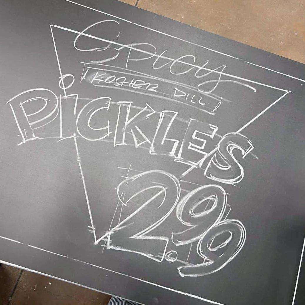 Trader Joe's hand-lettered chalkboard sign for Spicy Kosher Dill Pickles, $2.99. Price numerals oversized inside a triangle frame.