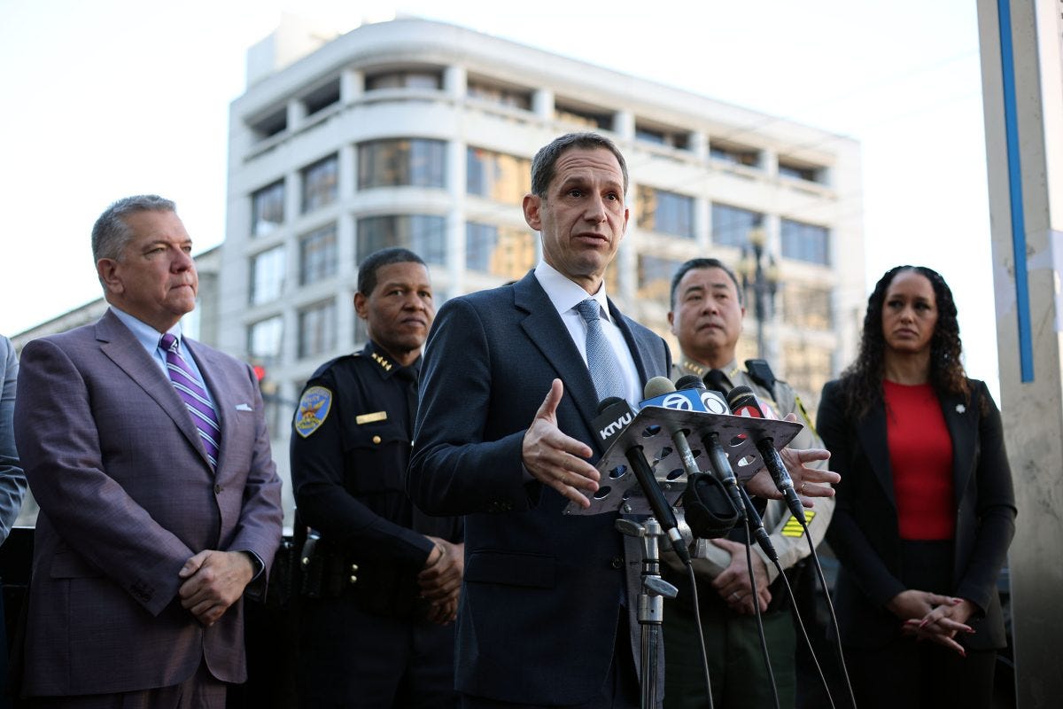A man in a suit speaks at a podium with microphones, flanked by four individuals. A city building is in the background. A man in a suit speaks at a podium with microphones, flanked by four individuals. A city building is in the background.