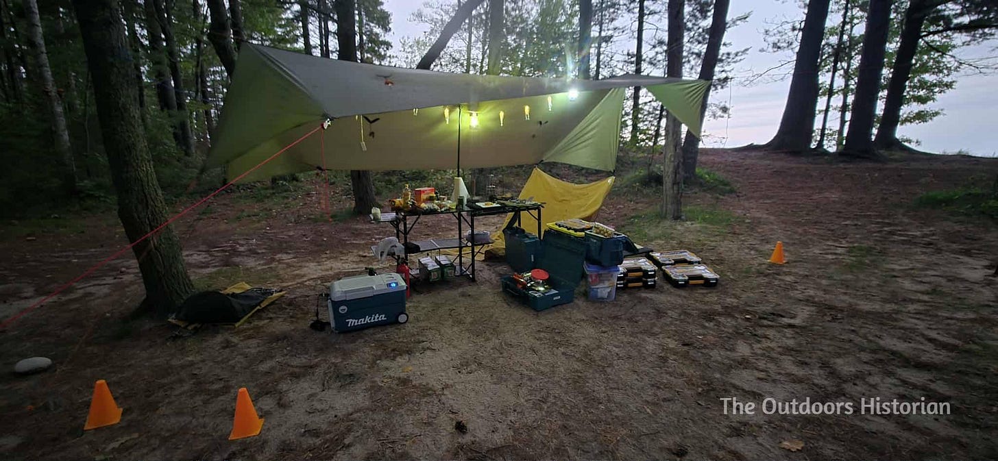 A tarp with a string of small skeleton lights along the ridgeline over a folding camping kitchen with a Makita cooler on the ground.