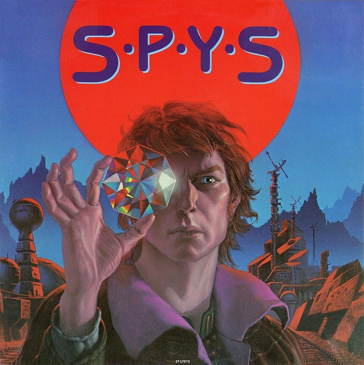 Front and back jacket illustration presented side by side. On the left, S•P•Y•S fills the bloody sun hanging in the sky. On the right, wraparound cover art further designs the scene with a squared arch in ruins as a succession of orbs trail across the sky, the structures that line the street are broken and a rundown building on the right is in decay.