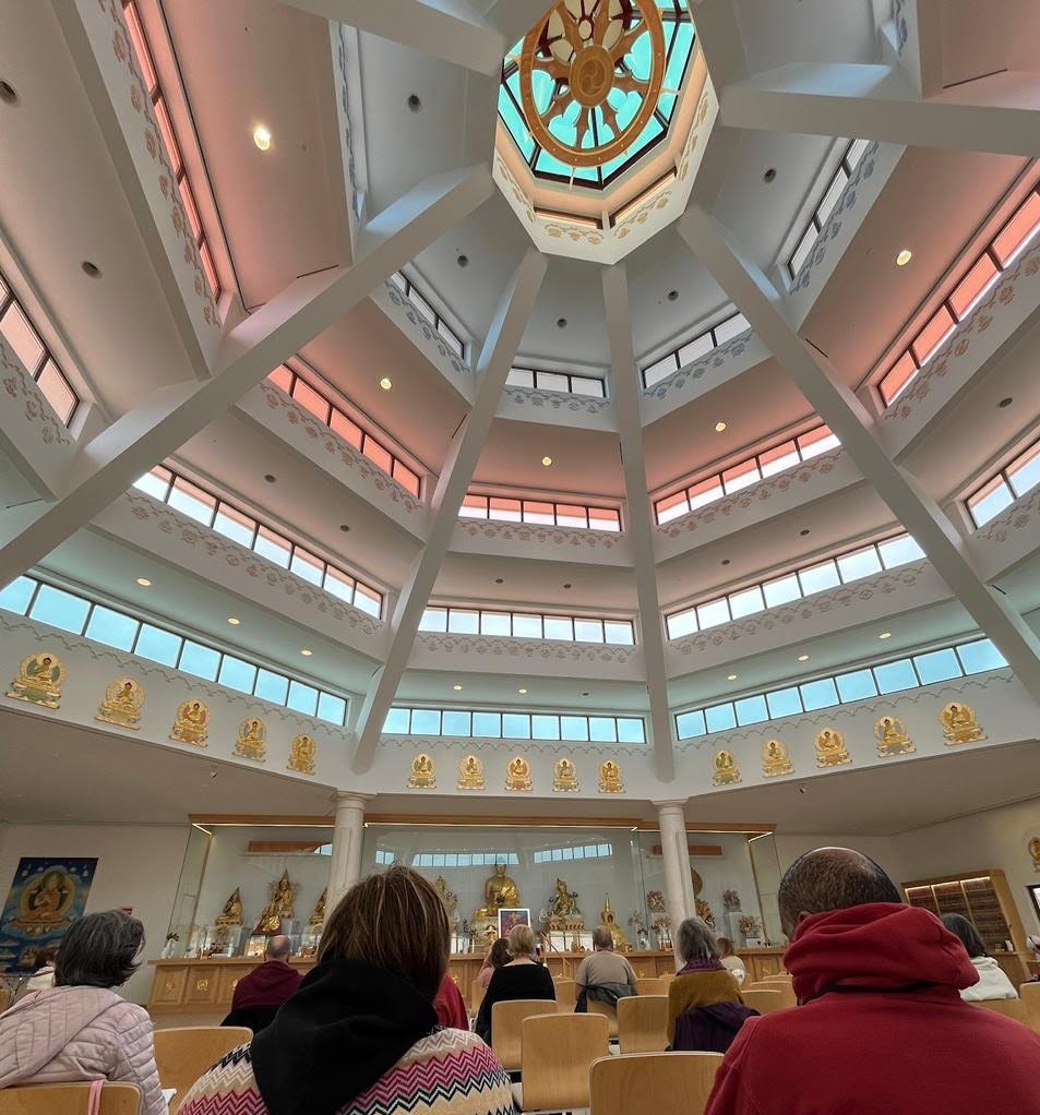 Inside the Kadampa meditation temple, attendees seated facing golden Buddha statues beneath an ornate octagonal skylight with teal and copper tones. Inside the Kadampa meditation temple, attendees seated facing golden Buddha statues beneath an ornate octagonal skylight with teal and copper tones.