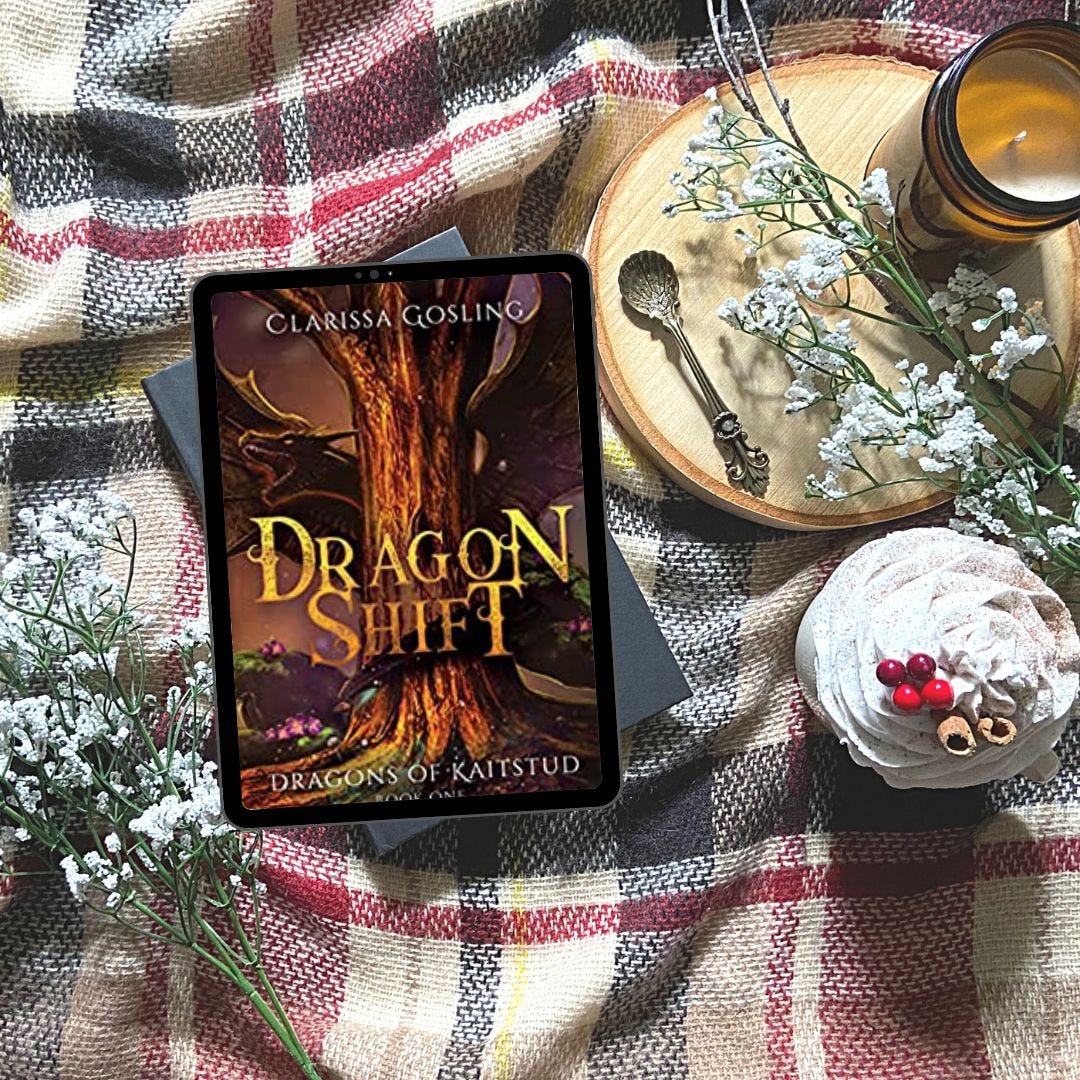 electronic tablet showing the cover of Dragon Shift by Clarissa Gosling lying on a plaid blanket with a cupcake, candle and flowers next to it