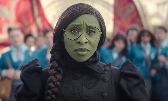 Cynthia Erivo with green makeup, braided hair, wearing round glasses and a decorative black dress, stands in front of a group in matching blue uniforms