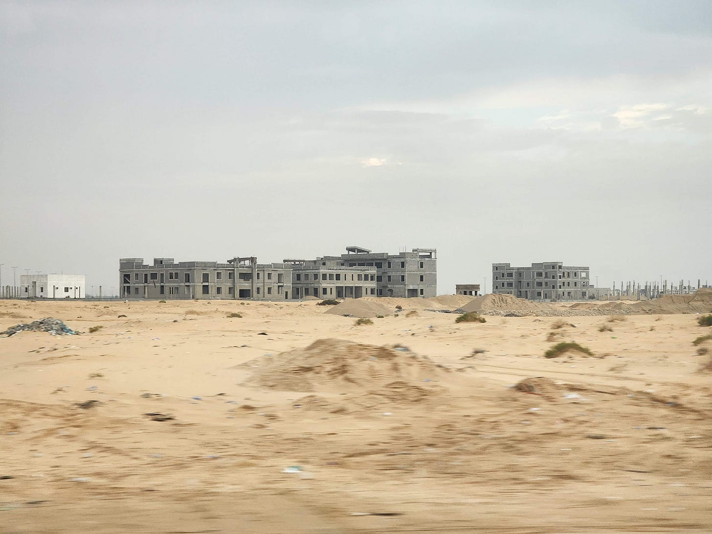Buildings under construction across a patch of sand, with piles of refuse in between.