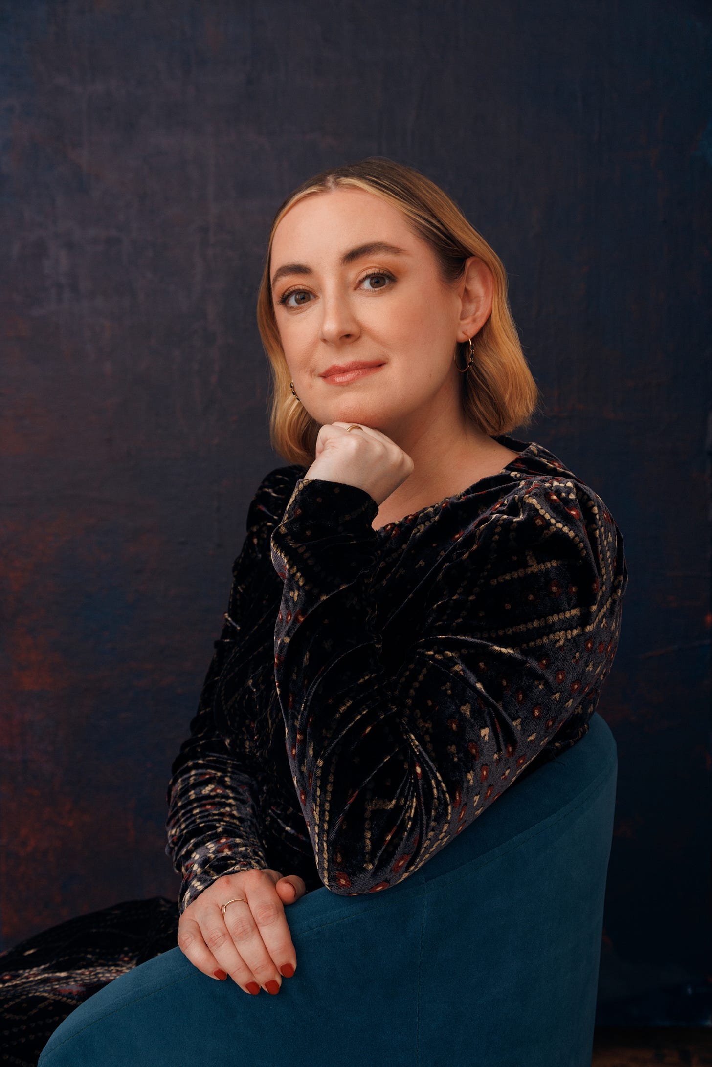 Author photo of Leigh Stein, sitting in a blue armchair and resting her chin in her hand