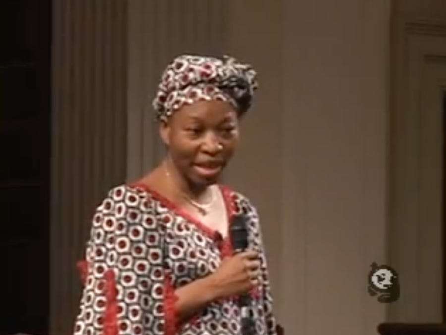An African woman wearing a dress and matching head wrap in a fabric patterned with small hexagons stands with a microphone, talking. An African woman wearing a dress and matching head wrap in a fabric patterned with small hexagons stands with a microphone, talking.
