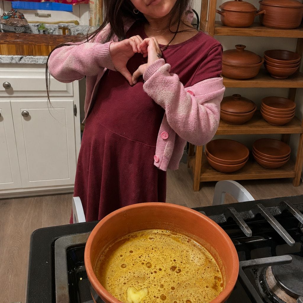 A young child in a burgundy dress and pink cardigan makes a heart shape with her hands behind a clay pot simmering on a stovetop, with shelves of earthen cookware in the background.