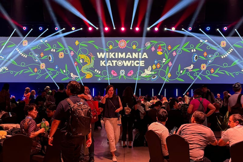 Large conference hall with crowd assembled around tables, room darkeneed with red and blue spotlight beams shining, "Wikimania Katowice" on wide projector screen in front