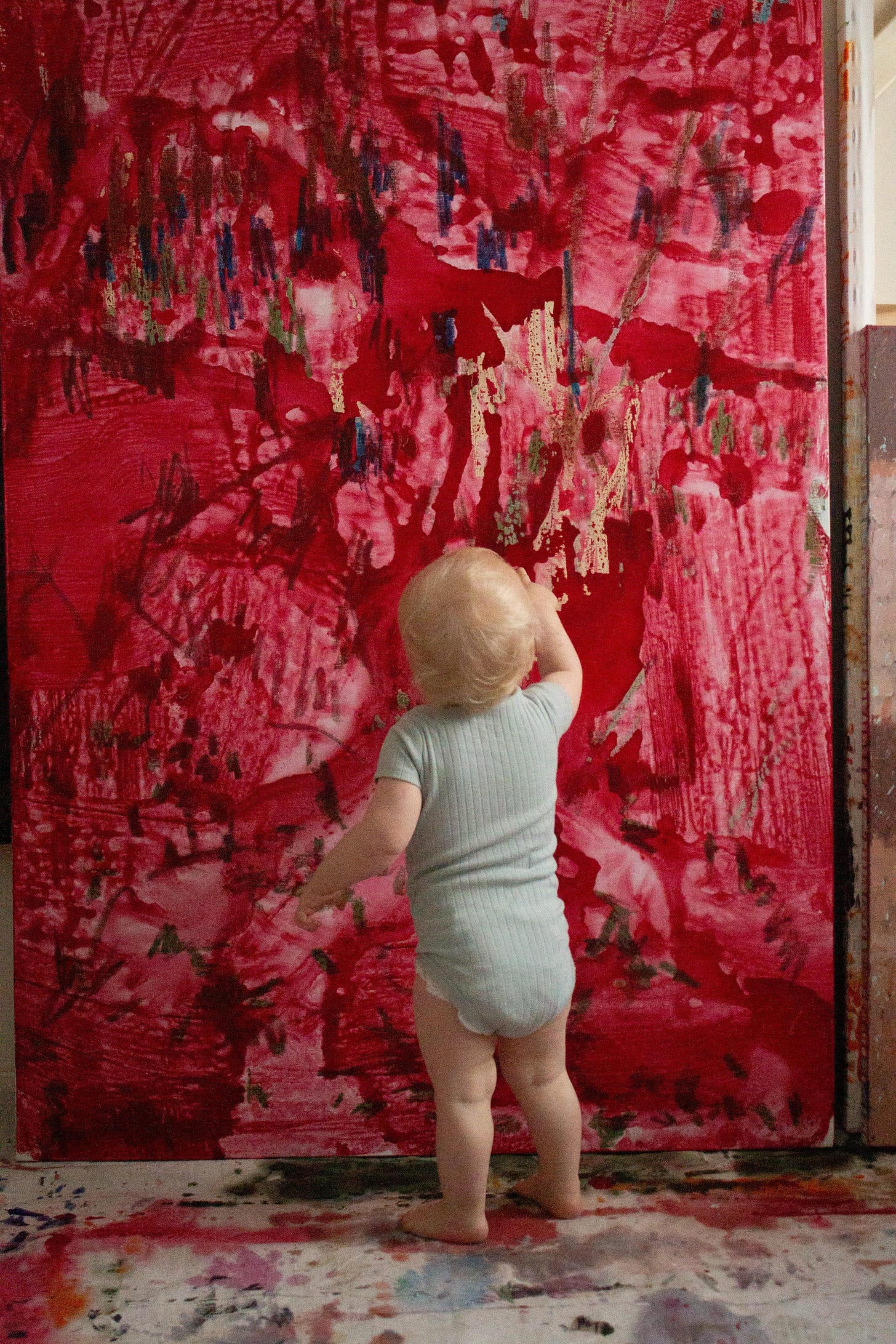  Infant standing facing a large red painting with a finger pointed up touching the surface.