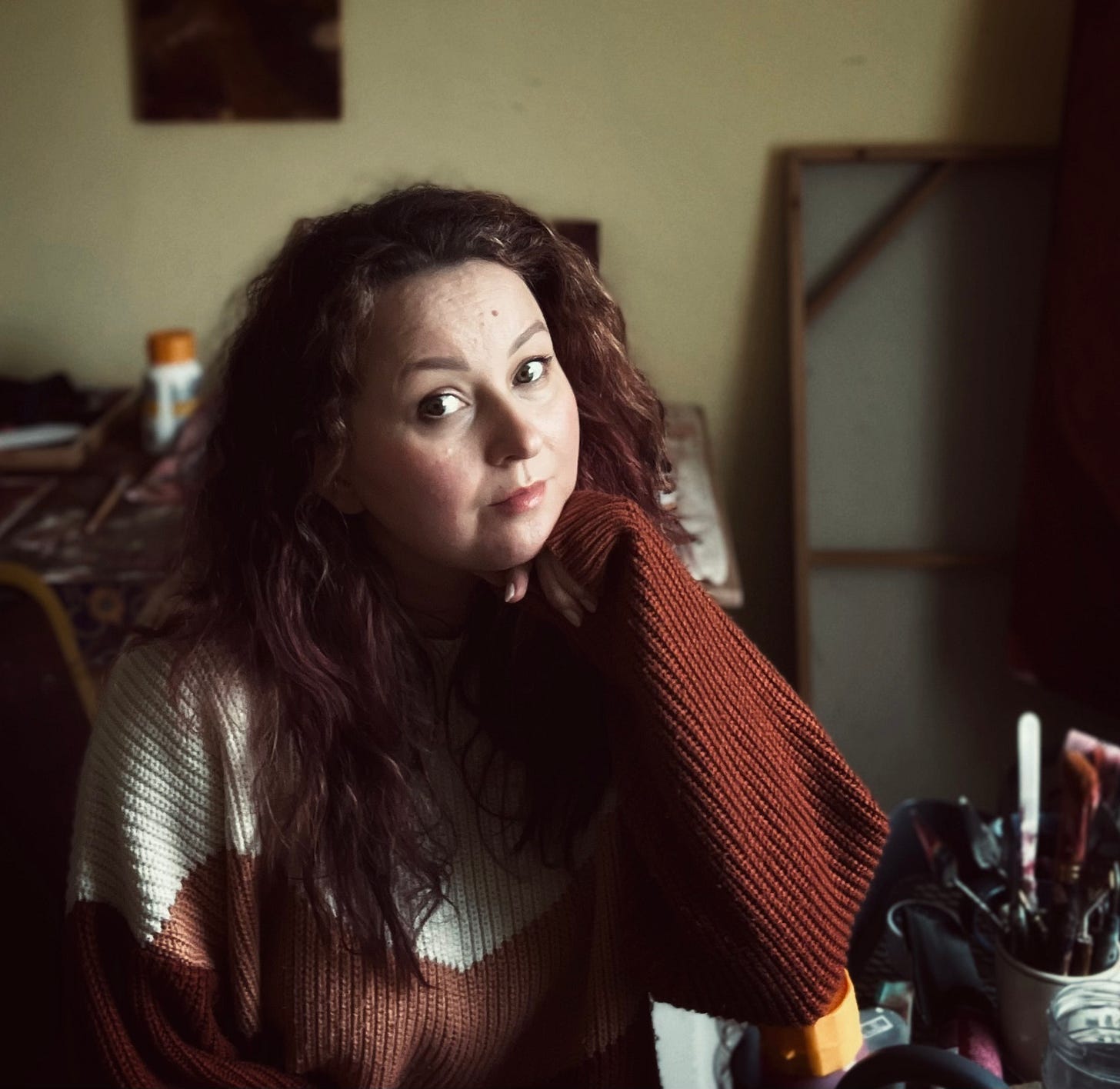 A softly lit portrait of the artist seated in her studio, wearing a warm rust and cream jumper. She rests her chin gently on her hand, looking out with calm, thoughtful presence.