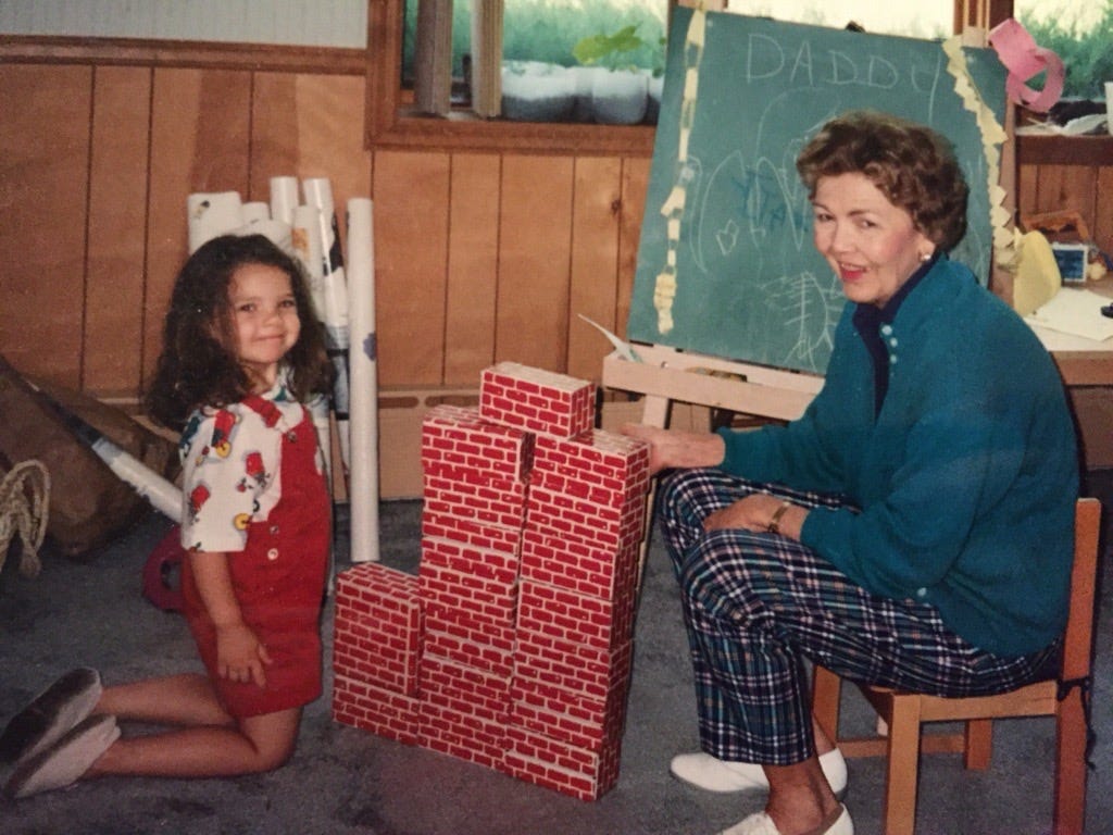 An image of a little girl in red overalls and a white teeshirt in a playroom next to a small tower constructed with fake bricks. Behind the tower is a chalkboard with a child's writing on it, and an older woman, who is the child's grandmother. Both the child and the grandmother are smiling.