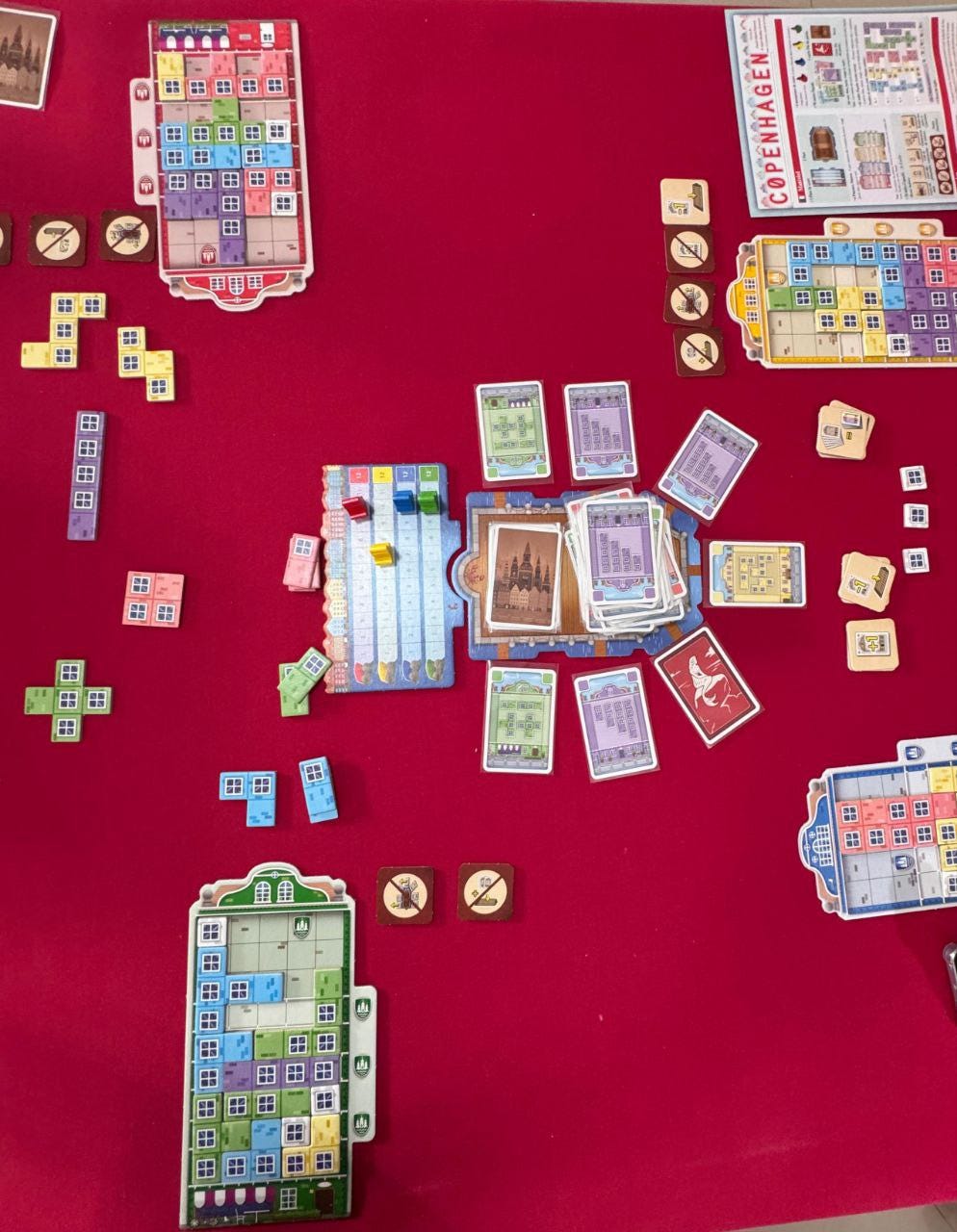 Foto de uma mesa com toalha vermelha. Ao centro há um pequeno tabuleiro com trilha de pontos e quatro peões. Os vermelho, azul e verde estão à frente, empatados, e o amarelo um pouco atrás. Também há um monte de cartas roxas, verdes, amarelas e vermelhas. Cada jogador tem um tabuleiro próprio com peças no estilo Tetris formando janelas de um edifício.