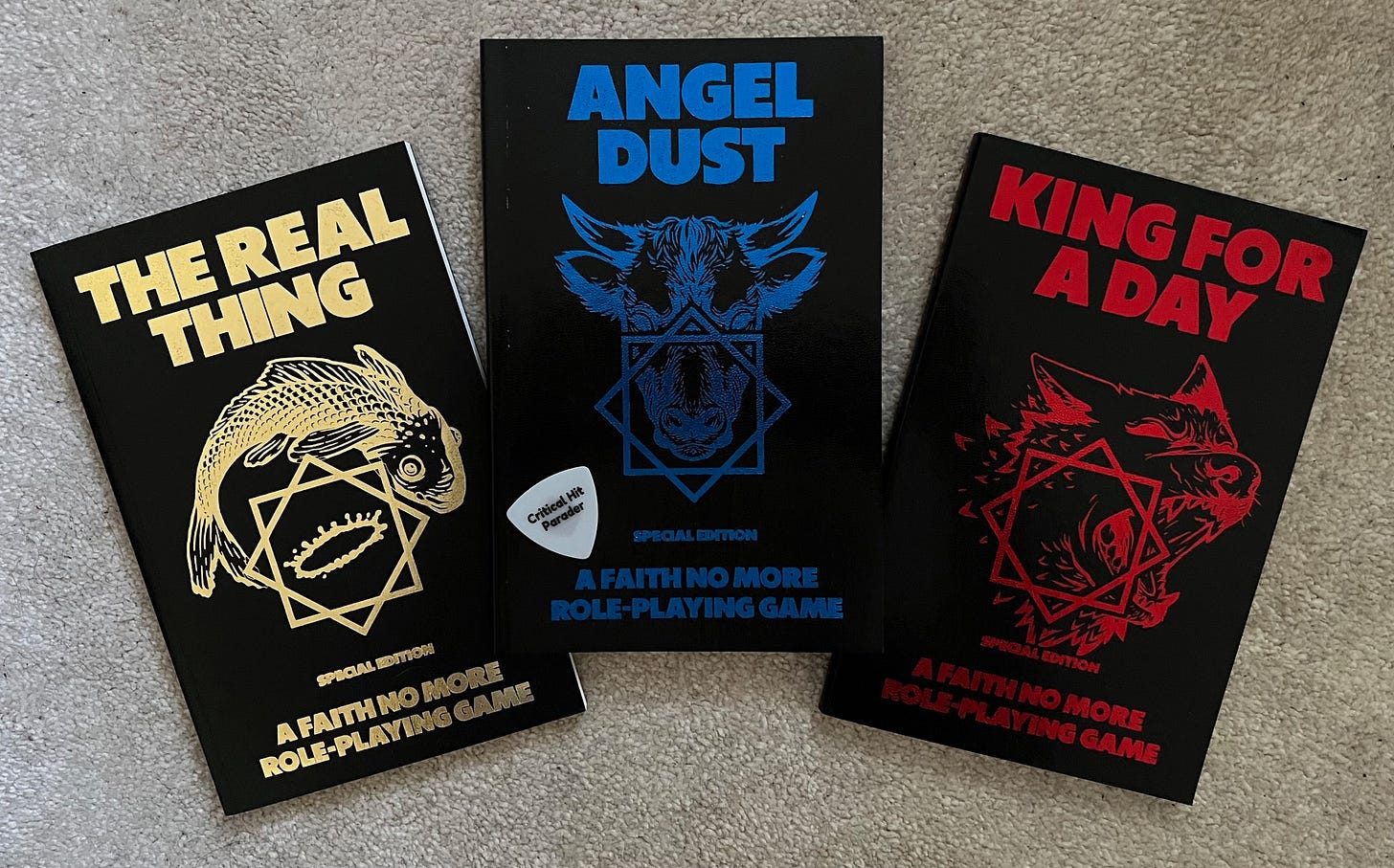 Three paperback books that make up the Faith No More Role-Playing Game. They all have black backgrounds. "The Real Thing" has gold text and a gold fish and crown symbol. "Angel Dust" has blue lettering and a bull symbol. "King for a Day" has red lettering.