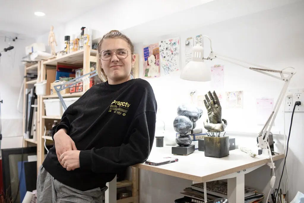 Aidan, wearing a black sweater, is leaning on a white art table, on which are several small metal sculptures. Paper works hang on the white walls and in the background, a tall wooden case holds various art supplies. Aidan, wearing a black sweater, is leaning on a white art table, on which are several small metal sculptures. Paper works hang on the white walls and in the background, a tall wooden case holds various art supplies.