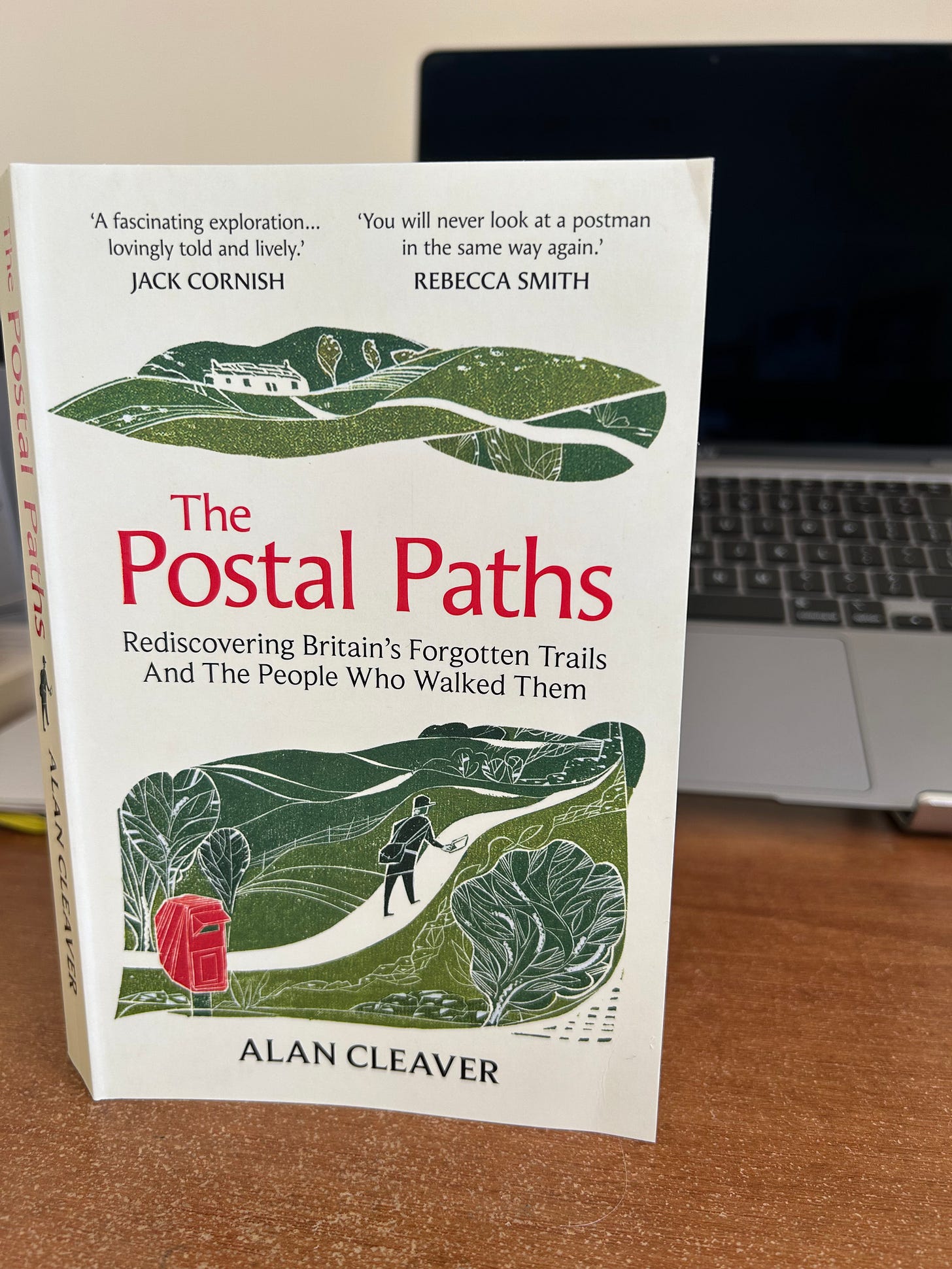The book, "The Postal Paths" by Alan Cleaver stands end on in front ot an Apple Macbook Air on a desk.