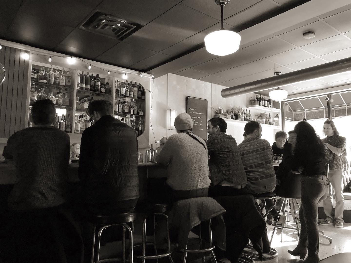 A black and white photo of several people sitting at a bar. A black and white photo of several people sitting at a bar.