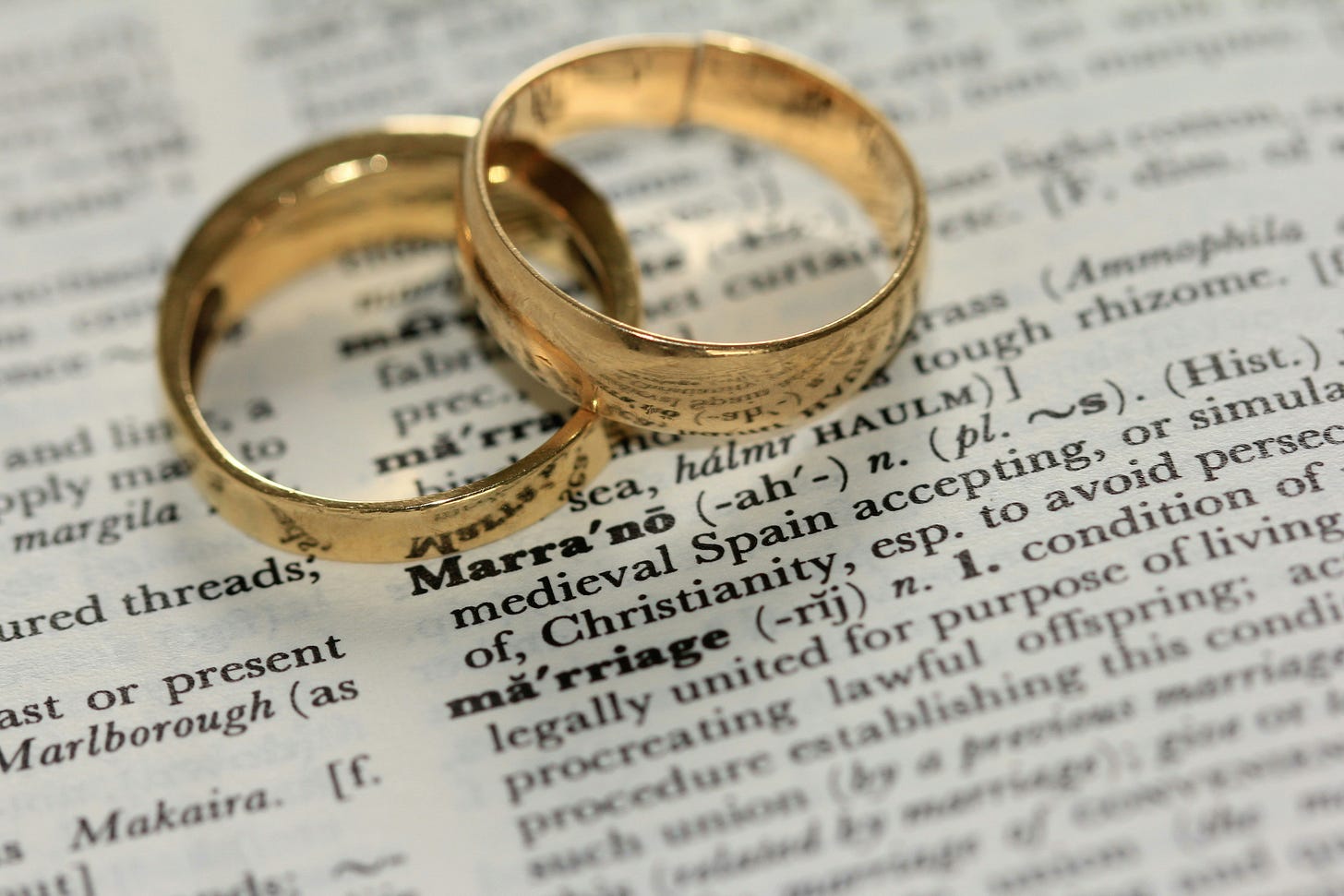 Two gold wedding rings resting on a dictionary page open to the definition of “marriage.”