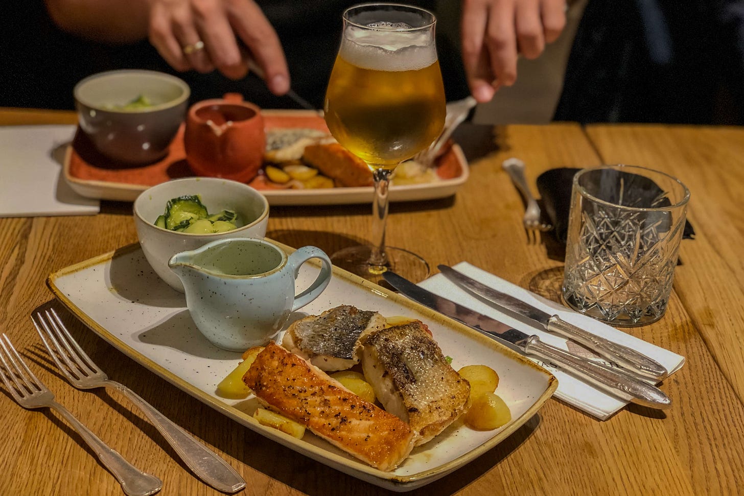 Gedeckter Holztisch im Restaurant mit einem Teller gebratenem Fisch auf Kartoffeln, dazu Gurkensalat und eine kleine Sauciere; daneben Besteck, ein Wasserglas und ein Glas helles Bier, im Hintergrund schneiden Hände weiteres Essen auf einem zweiten Teller. Gedeckter Holztisch im Restaurant mit einem Teller gebratenem Fisch auf Kartoffeln, dazu Gurkensalat und eine kleine Sauciere; daneben Besteck, ein Wasserglas und ein Glas helles Bier, im Hintergrund schneiden Hände weiteres Essen auf einem zweiten Teller.
