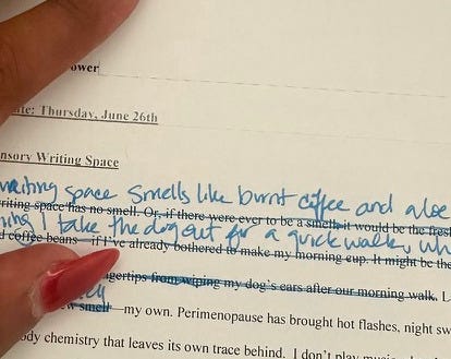 a brown hand with red fingernails holds down a piece of paper with printed text in black and handwritten revisions above the text in blue ink. 