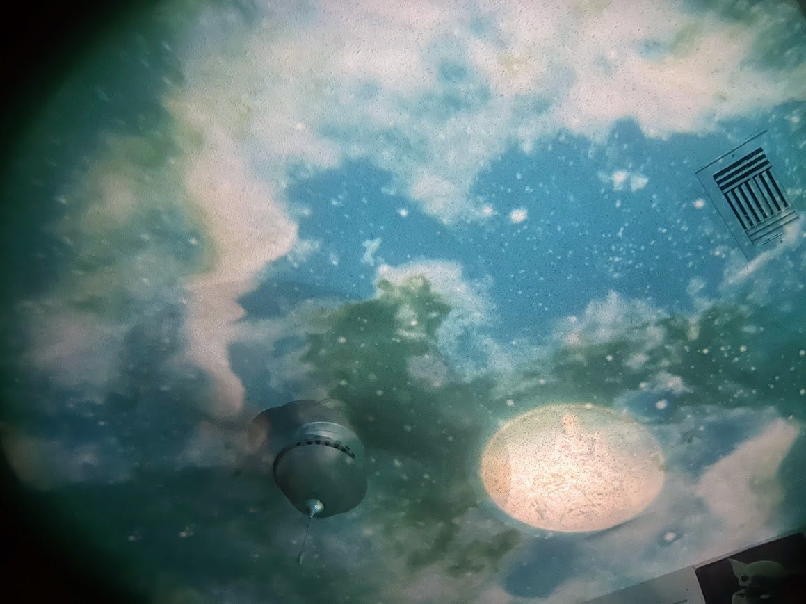 Photo of a bedroom ceiling in the dark illuminated with a space-themed scene including hazy white parts, a moon, and stars.