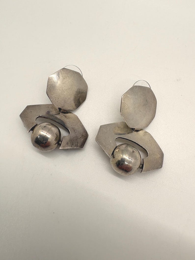 May include: A pair of silver-toned earrings. Each earring features a geometric design with a large, round ball and a flat, octagonal top. The earrings have a vintage or retro aesthetic.