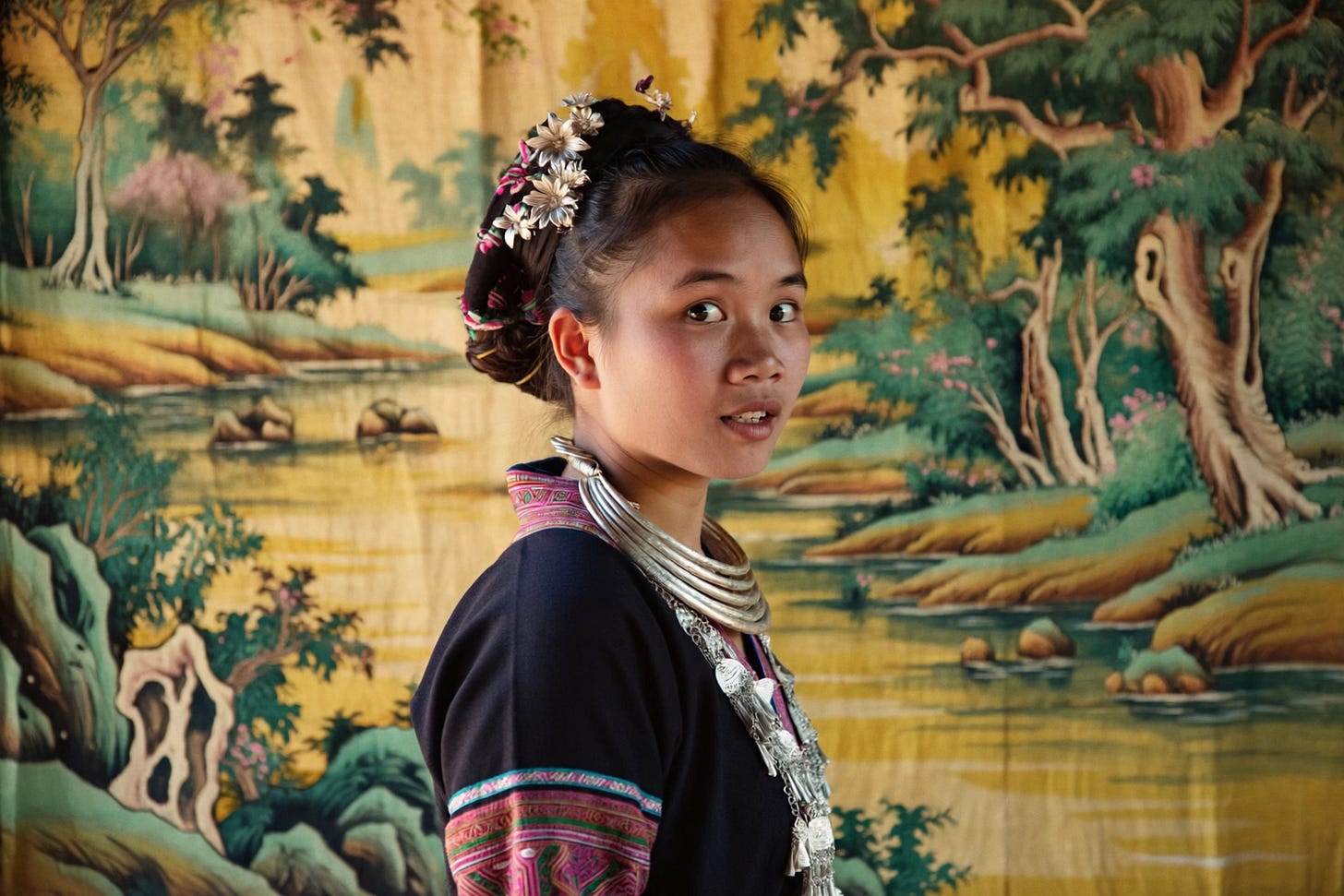 Candid photograph of a young Vietnamese Hmong woman wearing traditional jewelry, clothing, and hair styling; heavy, layered silver necklaces and intricate floral hairpieces. Her body is half-turned away, and she is looking back at the camera as if surprised. The background is a traditional Vietnamese silk painting showing a river and trees, in yellow and green with pink highlights. Dutch angle, three-quarter portrait.