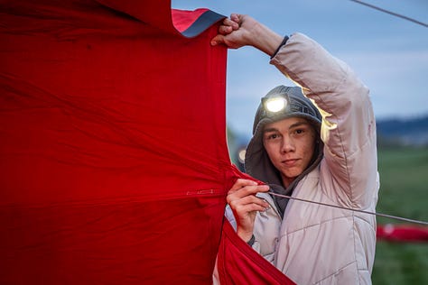 Înainte ca baloanele să urce, totul începe jos, pe iarbă, în frigul subțire al dimineții sau în lumina care abia se stinge peste câmp. Oamenii își ocupă locurile, verifică arzătoarele, întind pânza, ridică nacela, privesc cerul și, mai ales, ascultă vântul. În jur, mașinile stau cu farurile aprinse.