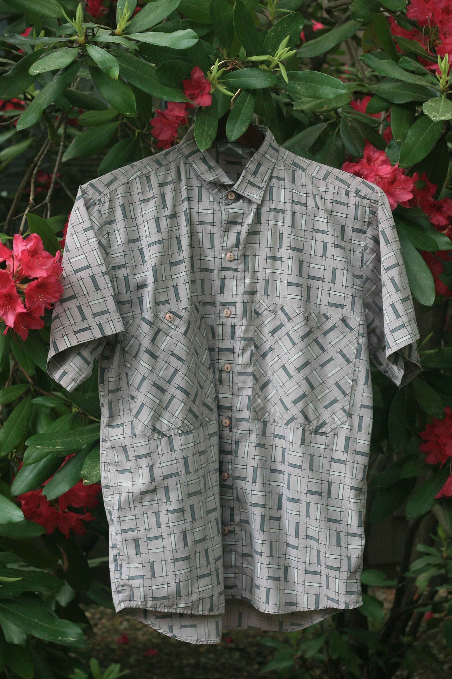 A short-sleeved, button-up shirt from the mid-1980s hangs on a wooden hanger among green leaves and bright red flowers. The shirt is light gray with a teal-green geometric grid pattern, two large chest pockets cut on the diagonal, and small beige buttons.