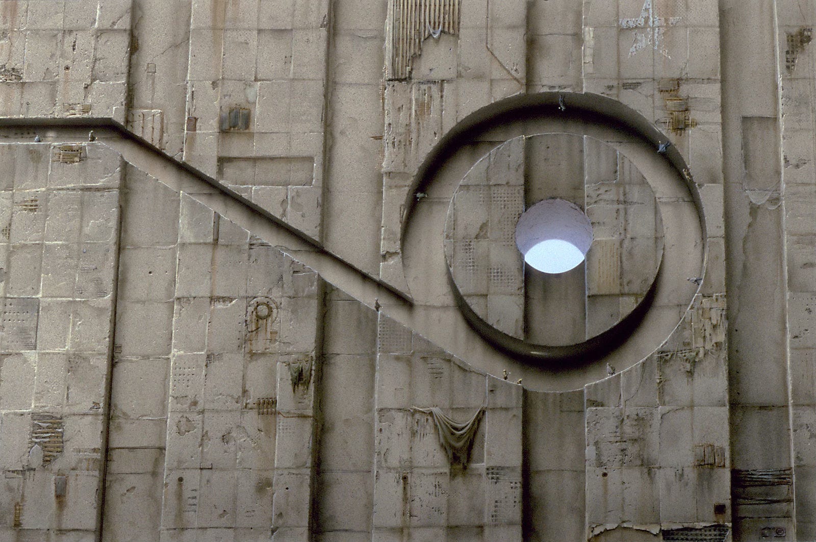 Tiny figures, each carrying a light, walk a path cut into the face of an enormous textured stone facade. The path angles down before opening to a round circuit where people seem to be defying gravity as they walk the curve even upside down. The channel in the stone resembles the number 9 lying face up. A hole at the center of the loop exposes the sky outside. A figure points his light up toward it but there is no apparent path  to get there. On the upper edge of the panel, a weathered five-pointed star is painted on the stone in white.