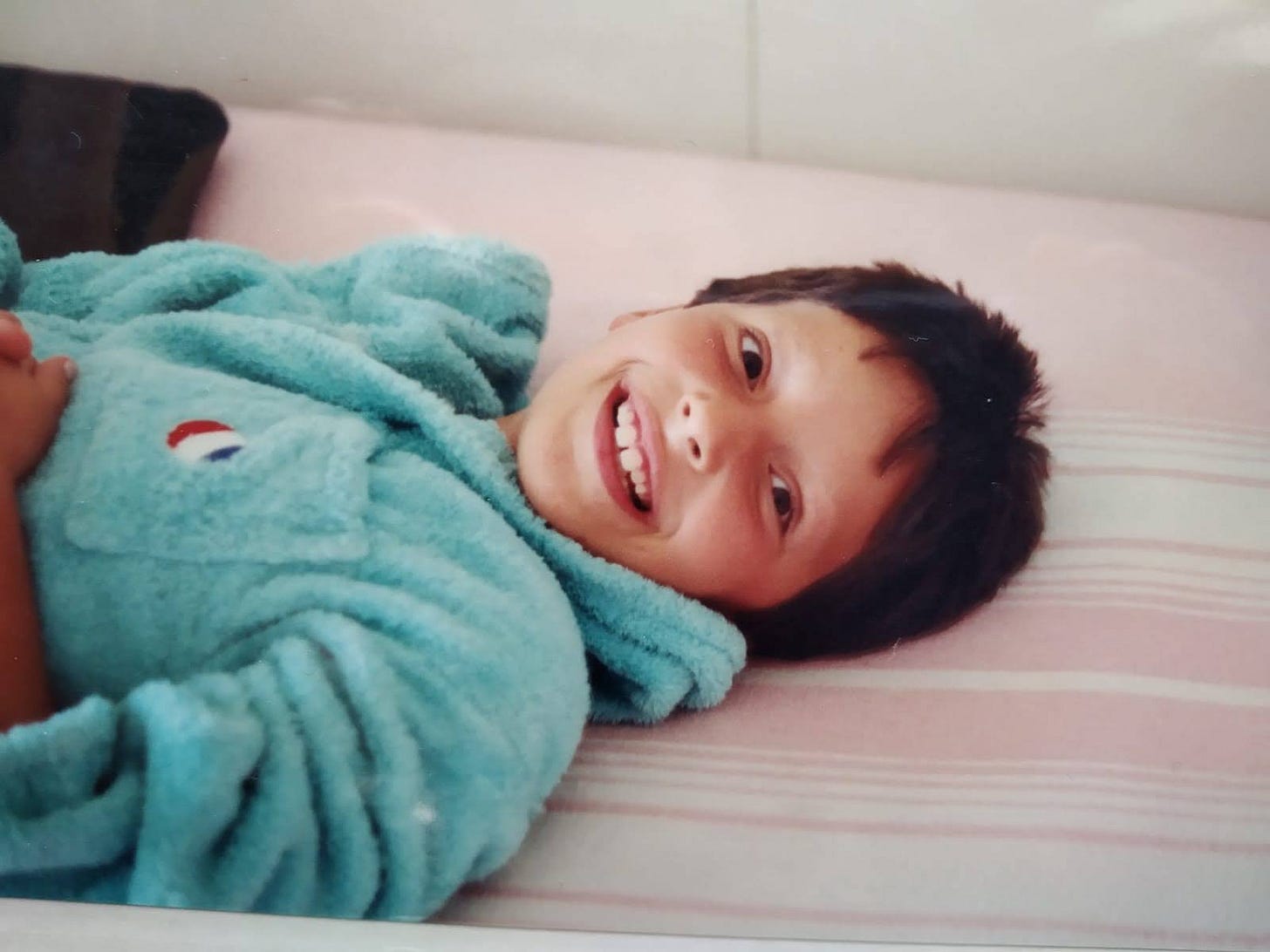 Bambina sdraiata, sorridente, con i capelli corti. Indossa un accappatoio verde mare, guarda in camera. Bambina sdraiata, sorridente, con i capelli corti. Indossa un accappatoio verde mare, guarda in camera.