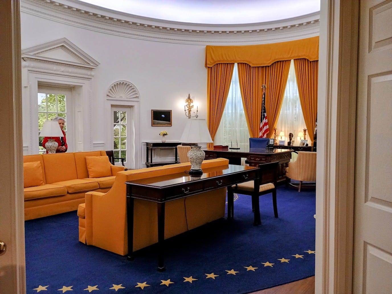 Reproduction of Oval office with gold curtains and sofas, woman partly hidden by lampshade