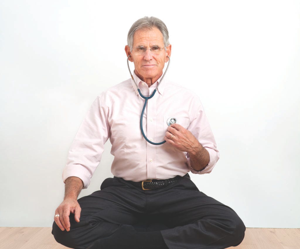 Man in pink shirt and black pants uses stethoscope, sitting cross-legged on light wood floor. Background is plain white.