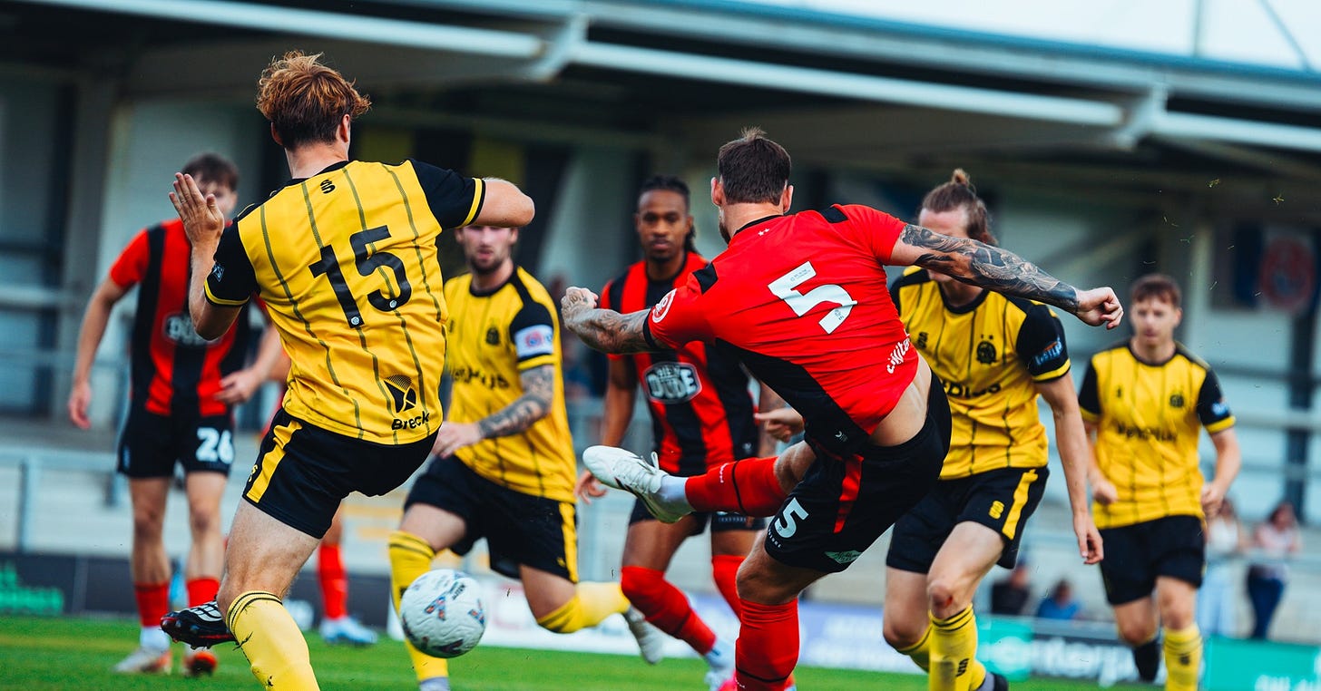 Number 5 in red (Huddersfield Town) strikes the ball on the edge of the box as a Fylde defender (yellow) attempts to block, with other players from both sides in the area. Number 5 in red (Huddersfield Town) strikes the ball on the edge of the box as a Fylde defender (yellow) attempts to block, with other players from both sides in the area.