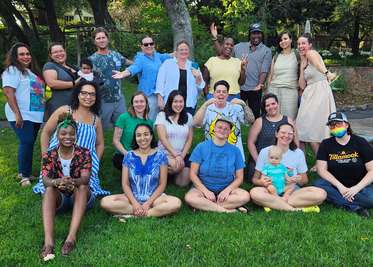 Group of 19 adults and 2 babies smiling for a group photo while sitting and standing on a lawn, outside under some trees. All are smiling, some are holding their hands out.