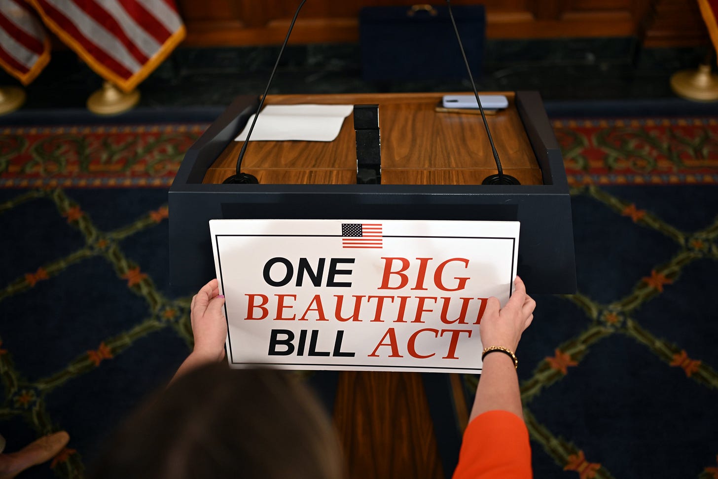 A white woman places a sign One Big Beautiful Bill Act in red and blue capital letters on a podium.