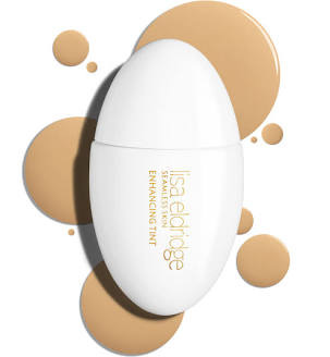 An elegant, matte white, slightly curved bottle with a simple gold font reading 'Lisa Eldridge Seamless Skin Enhancing Tint'. It's positioned next to large, textured dollops and swatches of the foundation in a warm, 'drippy' golden-light shade. An elegant, matte white, slightly curved bottle with a simple gold font reading 'Lisa Eldridge Seamless Skin Enhancing Tint'. It's positioned next to large, textured dollops and swatches of the foundation in a warm, 'drippy' golden-light shade.