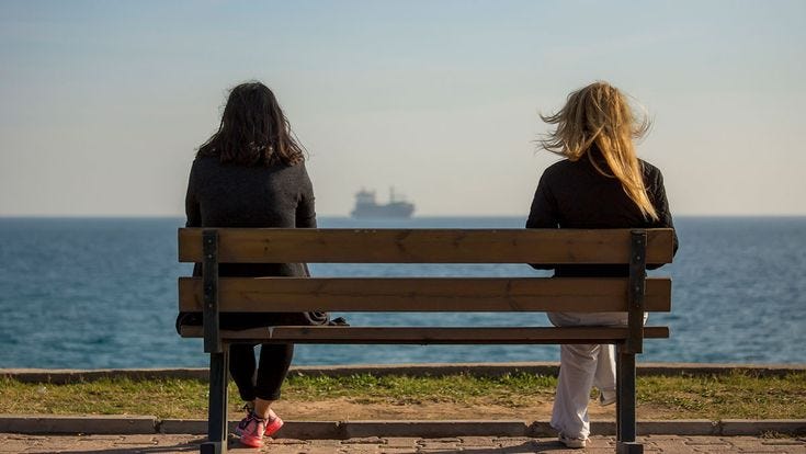 This may contain: two women sitting on a bench looking out at the ocean with a ship in the distance