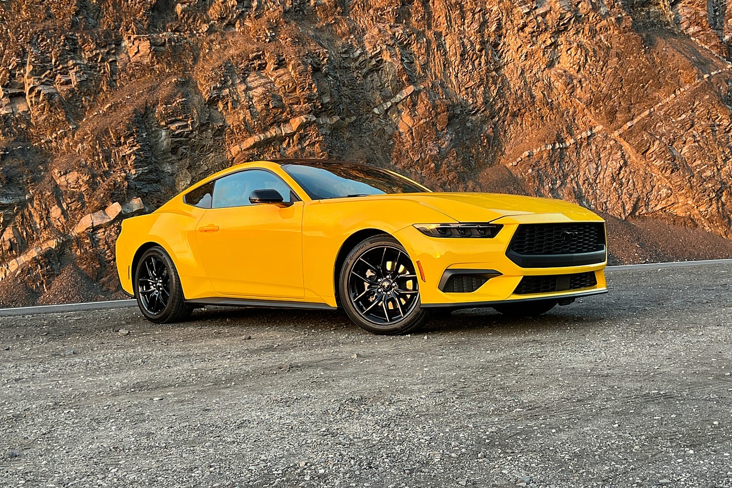 A yellow 2024 Ford Mustang coupe is parked on gravel with a rock wall behind it, lit by the sunset.