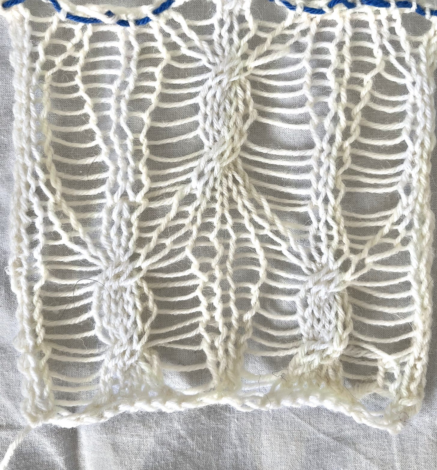 A small unblocked hand-knitted swatch in cream yarn showing the same diamond lace pattern as the machine-knitted scarf. 