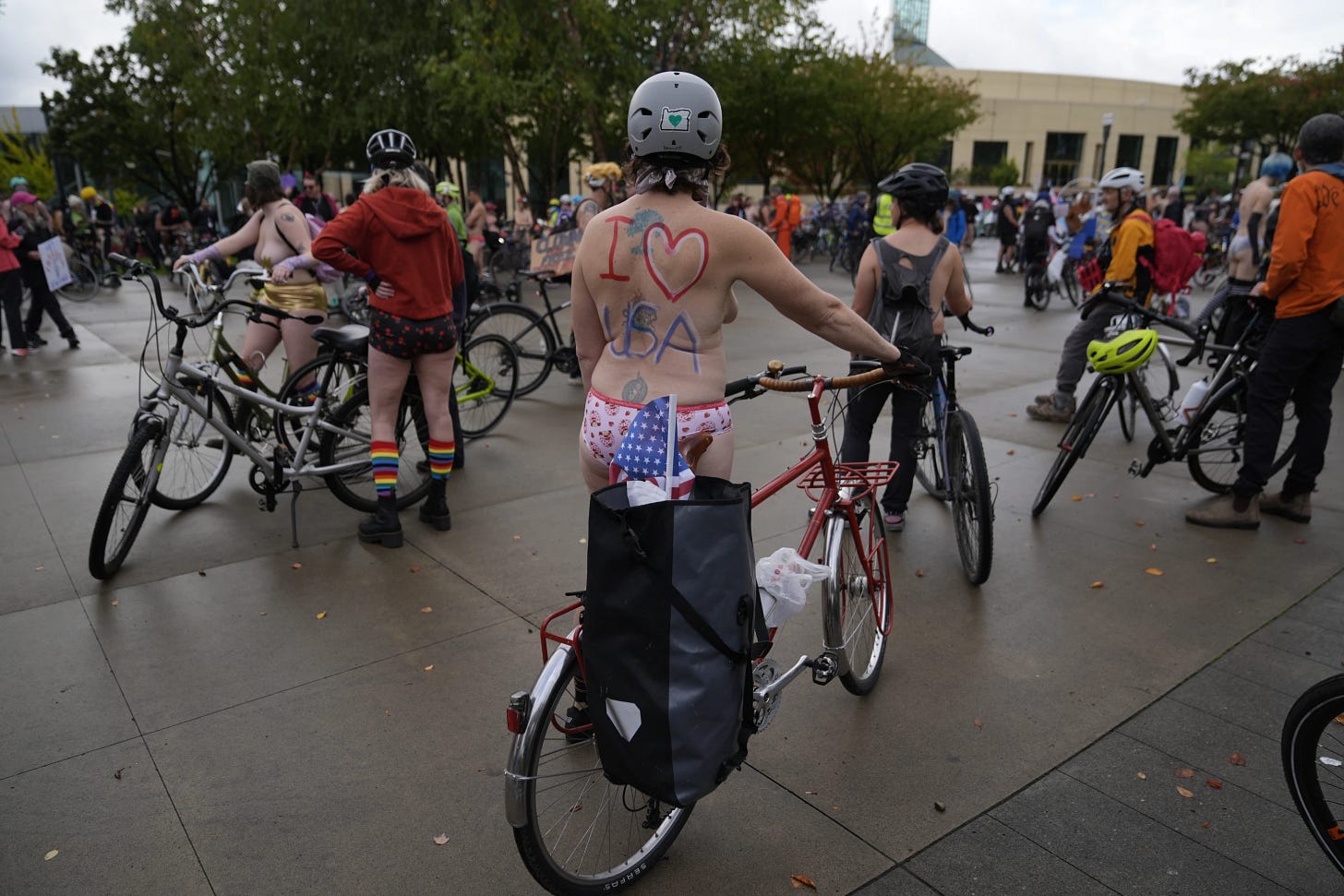 A partially nude cyclist with “I ❤️ USA” painted on their back stands beside a red bike among other riders gathering in a plaza before the protest ride in Portland. A partially nude cyclist with “I ❤️ USA” painted on their back stands beside a red bike among other riders gathering in a plaza before the protest ride in Portland.
