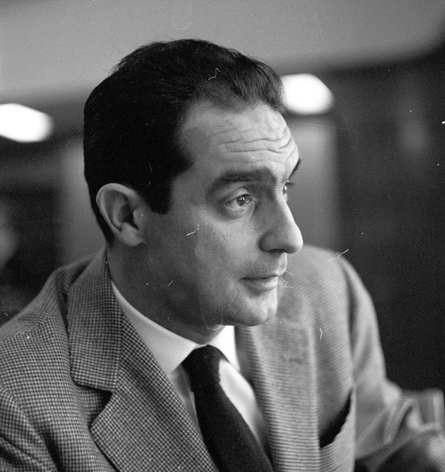 A man in a suit and tie is looking to the side in a black and white photo, with a blurred indoor background.